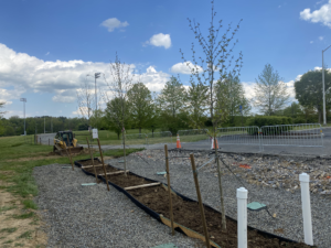 An installed multi-tree gravel bed stormwater retention system. The bed runs parallel to a parking lot, just inside a large turf field. A backhoe is visible in the background.